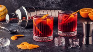 Zwei Negroni-Cocktails vor einem Shaker mit Eiswürfeln und Orangenzesten. 